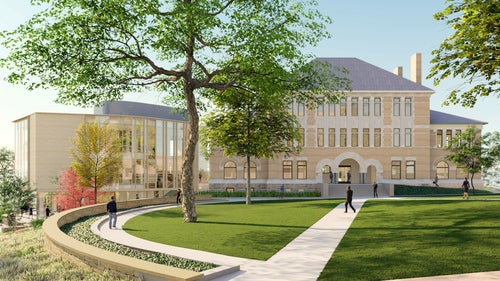 A large brick academic building with arched windows and a modern glass addition, surrounded by trees and landscaped lawns with people walking on pathways.
