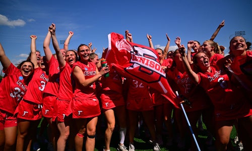 A photo of Denison's Women's Lacrosse team in uniform with a large Denison flag bearing the Block D Denison Athletics logo celebrates its Sweet Sixteen victory.