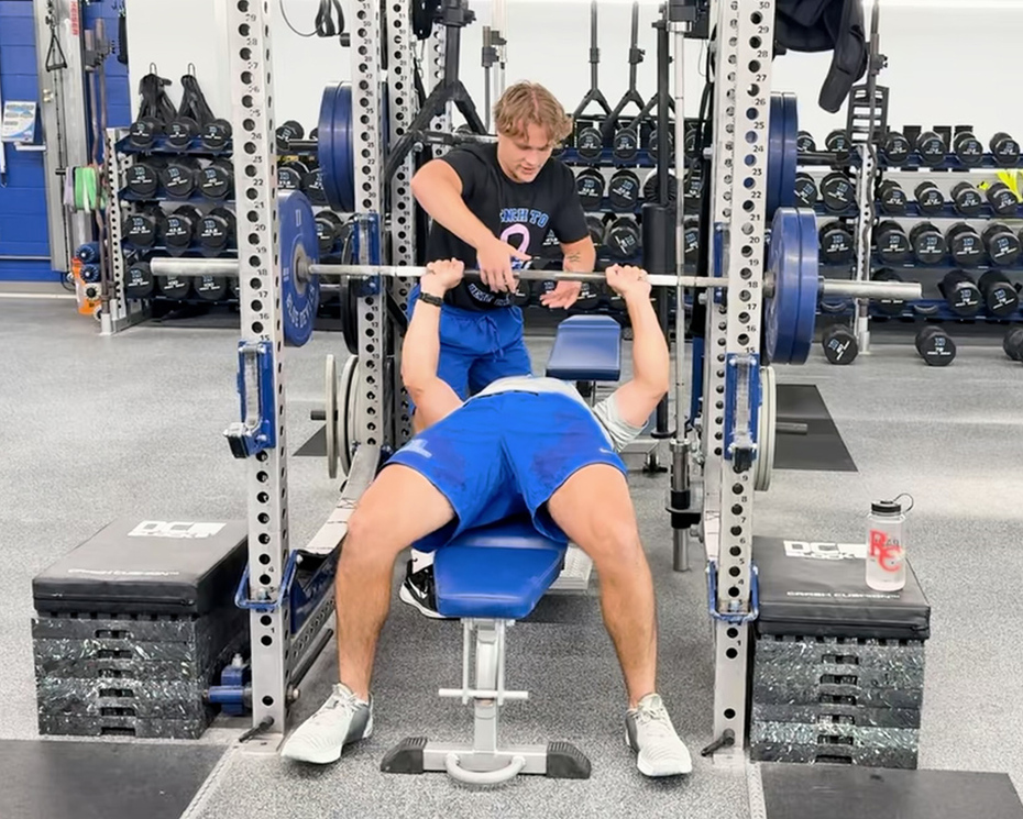 Denison student assists weightlifter with a rep on a weight bench in a gym.